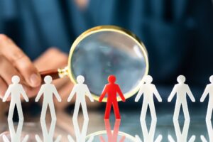Representation of hiring challenges - Line of white stick figures with magnifying class pointed at red one