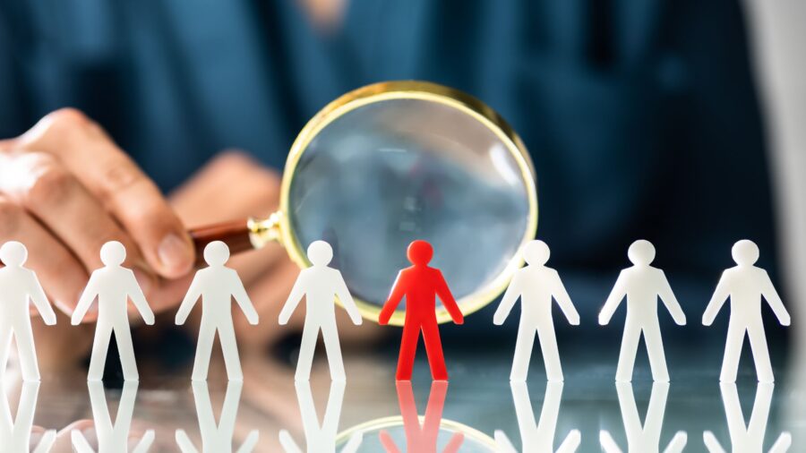 Representation of hiring challenges - Line of white stick figures with magnifying class pointed at red one